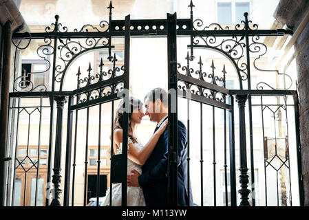 Horizontal close-up romantic portrait of the beautiful happy newlyweds rubbing noses behind the open irone fence door. Stock Photo
