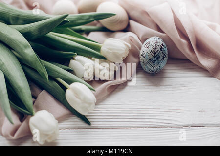 stylish white tulips and easter eggs with floral ornaments on rustic wooden background. happy easter greeting card concept with space for text. soft l Stock Photo