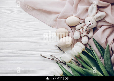 stylish easter eggs and willow buds and rabbit and white tulips on rustic wooden background and fabric flat lay. happy easter greeting card concept wi Stock Photo