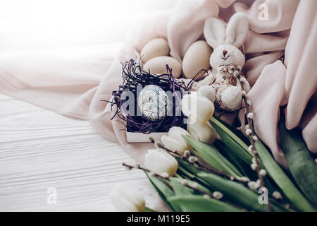 stylish easter eggs with chick ornaments in nest and bunny rabbit and tulips and willow buds on rustic white wooden background. happy easter greeting  Stock Photo