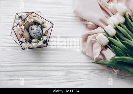 happy easter greeting card. stylish easter eggs with floral ornaments in nest of flowers and tulips on rustic white wooden background flat lay. space  Stock Photo