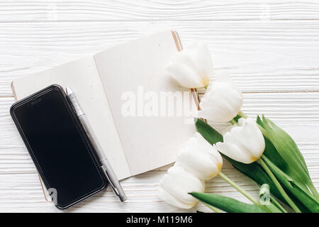 phone screen and notebook pen and stylish tulips on white wooden rustic background. flat lay with flowers and paper note with space for text. woman wo Stock Photo