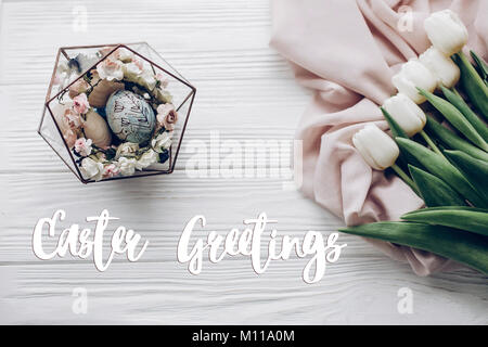 easter greetings text card sign on stylish easter eggs with floral ornaments in nest of flowers and tulips on rustic white wooden background flat lay. Stock Photo