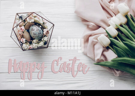 happy easter text greeting card sign on stylish easter eggs with floral ornaments in nest of flowers and tulips on rustic white wooden background flat Stock Photo