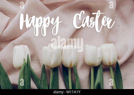happy easter text greeting card sign on spring flat lay. stylish white tulips on beige soft fabric on rustic  table background top view. soft light, t Stock Photo