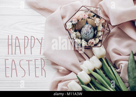 happy easter text greeting card sign on stylish easter egg with floral ornaments in nest of flowers and tulips on rustic white wooden background flat  Stock Photo