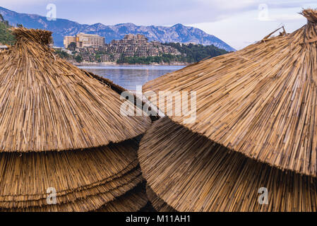 Straw parasols on a Slavic Beach in Budva city on the Adriatic Sea coast in Montenegro. Dukley Gardens Residential Complex on background Stock Photo