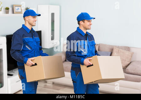 Young Smiling Professional Movers In Uniform Delivering Cardboard Boxes In Living Room Stock Photo