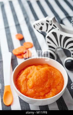 Pumpkin and carrot baby puree in bowl with baby spoon and zebra toy on striped tablecloth Stock Photo