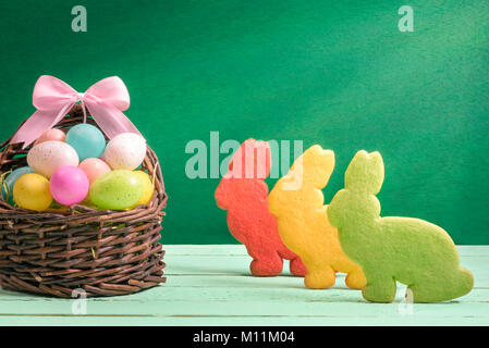 Easter banner design with a wicker basket full of colorful painted eggs, with a ribbon bow and three bunny shaped cookies, on a green background. Stock Photo