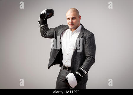 A young bald man in a white shirt, gray suit and boxing gloves seriously looks into the camera, shows a biceps and poses on a white isolated backgroun Stock Photo