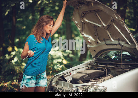 Beautiful young woman with a car that break down on the road in forest. She has standing by a car and holding wrench while looking at engine. Stock Photo