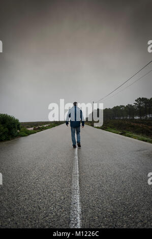 man walking down the middle of the road on a wet gloomy day. Back view. Stock Photo