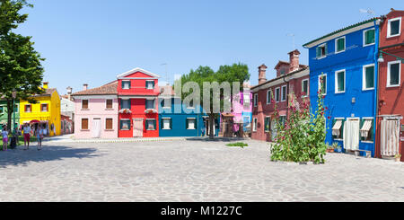 Panorama of brightly colored  houses in  Burano, Venice, Italy around a quiet square, Corte della Comare, with a couple walking through on a sunny day Stock Photo