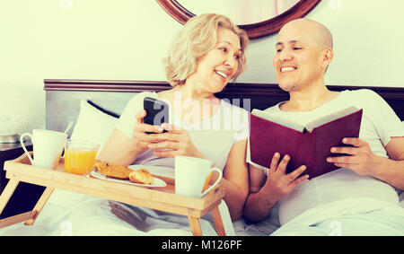 Cheerful elderly couple reading book while having breakfast in bed Stock Photo