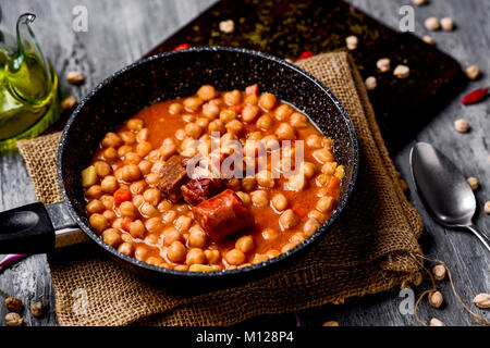 a stone frypan with garbanzos a la riojana, a spanish chickpeas stew, on a wooden chopping board placed on a rustic wooden table next to a cruet with  Stock Photo