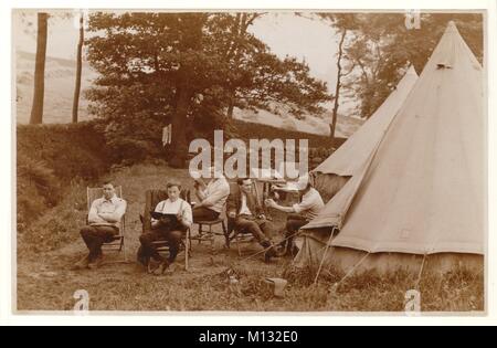 Early1900s post WW1 era postcard of young men camping, relaxing on chairs outside their canvas bell tents pitched in a field. Moorland in the background. One man is reading a book, one a newspaper and another pouring a cup of tea. Newly mown hay is drying in rows of stooks in the fields behind. Circa Summer 1920s, U.K. Stock Photo