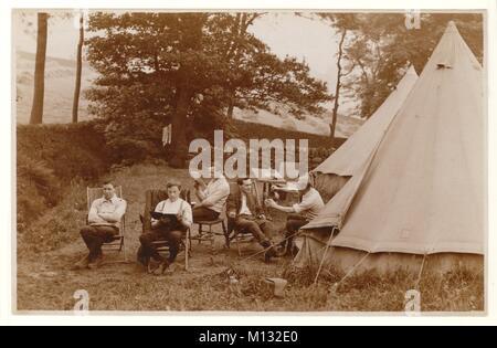 Early1900s postcard of young men camping, relaxing on chairs outside.  One man is reading a book, one a newspaper and another pouring a cup of tea. Newly mown hay is drying in rows of stooks in the fields behind. Circa Summer 1920s, U.K. Stock Photo