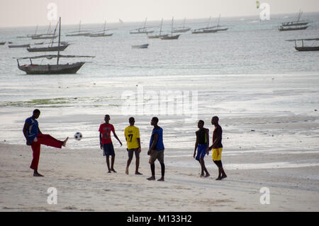 Local Tanzanian youths relaxing on the beach in Nungwi, Zanzibar, playing football in the setting sun with dhow boats in the background and soft light Stock Photo