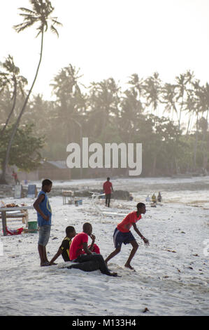 Local Tanzanian youths relaxing on the beach in Nungwi, Zanzibar, playing football in the setting sun with dhow boats in the background and soft light Stock Photo