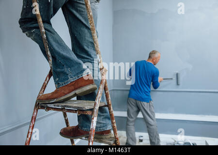 Two male builders are painting a room. One man is using a ladder and only his legs are visible. The other man is in the background. Stock Photo