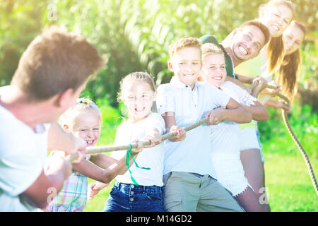 Kids with young moms and dads playing tug of war during joint outdoors games on sunny day Stock Photo