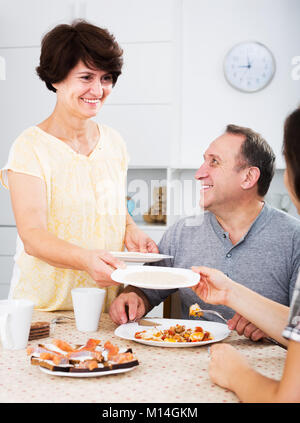 Smiling mature woman setting table for family lunch at home indoors. Focus on man Stock Photo