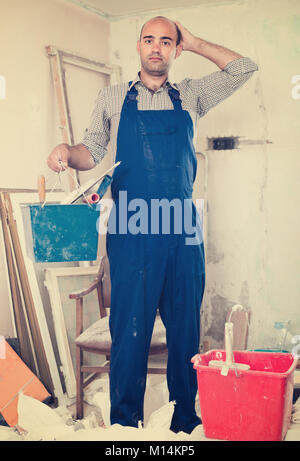 Builder is standing with bucket and putty knife and thinking about where he should start in uniform indoors. Stock Photo