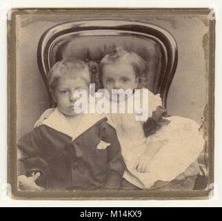 Charming original Victorian studio photographic portrait of a young boy with his baby sister, siblings, sitting in button-back armchair. The boy is wearing a fashionable sailor suit. Circa 1890s, U.K. Stock Photo