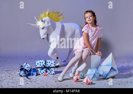 Fashion kid. Designer collection. White big unicorn origami made of paper. Girl in beautiful pink dress. Studio shot. Stock Photo