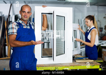 Cheerful workman in blue overalls demonstrating pvc window in workshop Stock Photo