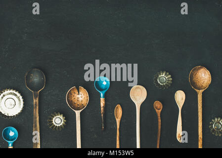 Flat-lay of various old vintage kitchen spoons and baking tin molds over black stone background, top view, copy space, horizontal composition. Rustic  Stock Photo