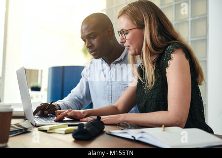 Two focused businesspeople sitting at a table in an office talking together and working online with a laptop Stock Photo