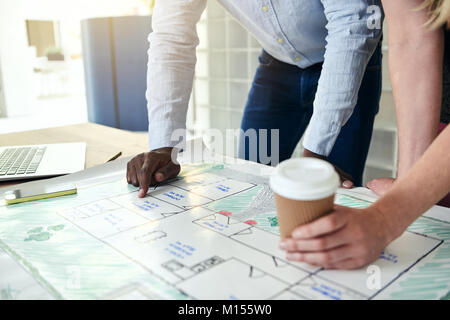 Two architects leaning on a table discussing blueprints and drinking coffee while working together in a modern office Stock Photo