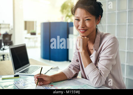Smiling young Asian designer sitting at a desk in a modern office looking over building designs Stock Photo
