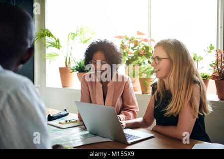 Two smiling female colleagues sitting together at a table in a boardroom talking with colleagues during a meeting Stock Photo