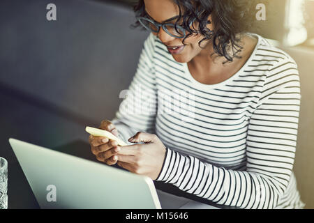 Young African woman wearing glasses sitting alone at a table sending a text message on her cellphone and working online with a laptop Stock Photo