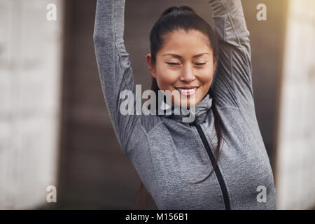 Young Asian woman in sportswear smiling while standing outside with her eyes closed and stretching her arms before exercising Stock Photo