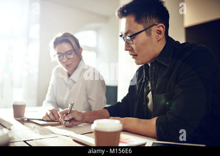 Two diverse work colleagues sitting together at a table in a modern office discussing paperwork Stock Photo