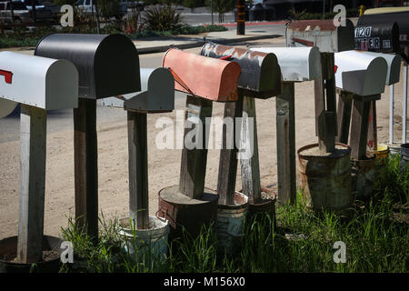 A row of many old metal mail boxes on top of wooden posts set into tubs of cement along a rural road, viewed from behind, some leaning, various colors Stock Photo