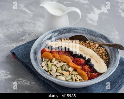 Muesli with banana, blueberries, dried apricots, carnelian cherry, peanuts and pumpkin seeds, in an old plate on a gray table Stock Photo