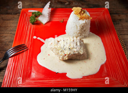 Steamed fillet of mahi-mahi fish in white sauce with rice and garlic chips served on a red square plate. Bali, Indonesia. Stock Photo