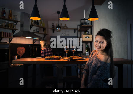 Young women preparing dinner in the evening. Blonde staying back on the background. Food and wine on the bar counter. Painted heart on the wall Stock Photo