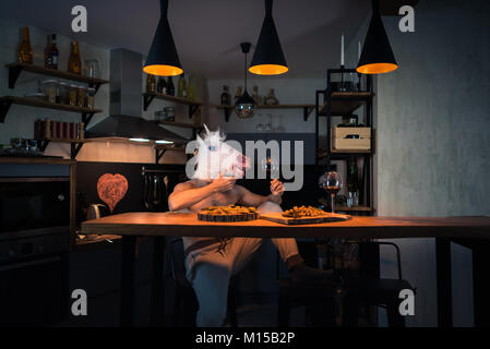 Funny unicorn sits alone at the bar counter in stylish apartment. Fantasy person. Young man in rubber mask at the table. Painted heart on the wall Stock Photo