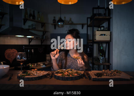 Young woman drinks wine and sits at the table with food. Romantic brunette alone dinning and dreaming in the evening. Painted heart on the wall Stock Photo