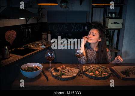 Young woman with closed eyes sits at the table with food and wine in the modern kitchen. Cute brunette alone dinning and dreaming in apartments Stock Photo