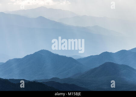 Scenic hazy blue mountains range covered by soft fog touching sunlight from cloudy sky. Stock Photo