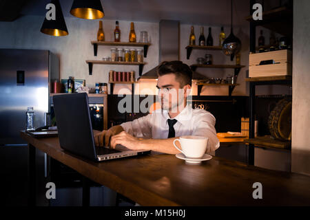 Young man sits in the kitchen at the bar counter and works in laptop. Home atmosphere, work in the evening with cup of coffee. Home office. Stock Photo