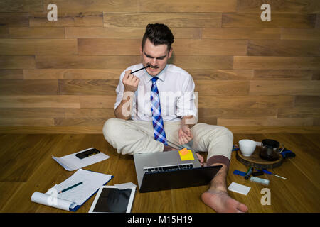 Young man works at home in funny clothes with a lot of gadgets and reads the news. Barefoot businessman sitting on the floor against a wall Stock Photo