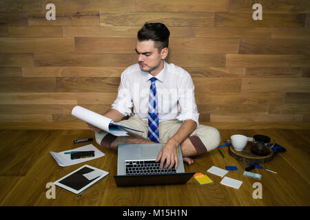 A barefoot businessman sits on the floor against a wall and using a laptop and other gadgets. Serious young man works at home in funny clothes Stock Photo